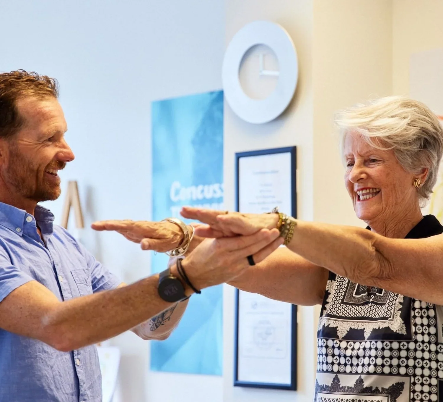 An elderly woman and a middle-aged man are smiling and playfully holding their hands out towards each other in a friendly greeting or game, standing indoors with a Paris poster and framed certificates on the wall.