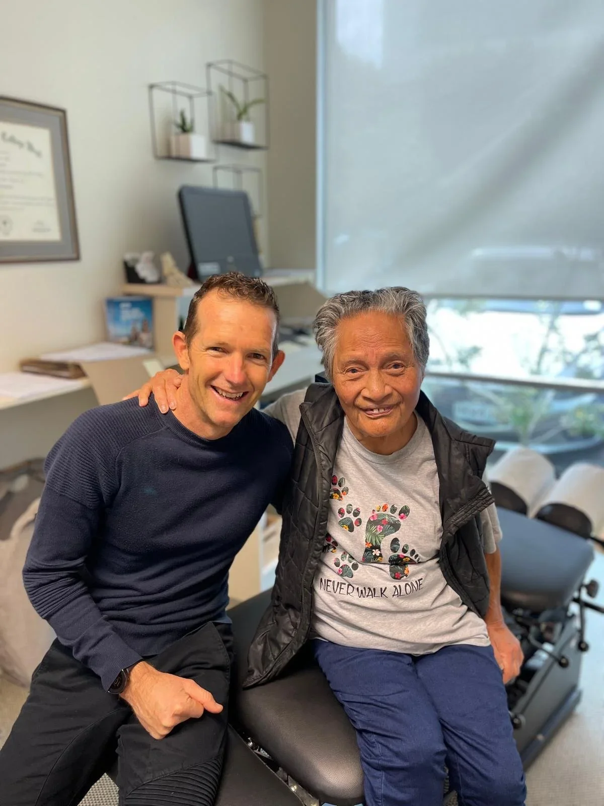 A young man and an elderly woman smiling together in a medical or therapy office, with the woman seated on a treatment table and the man standing beside her, both with arms around each other.