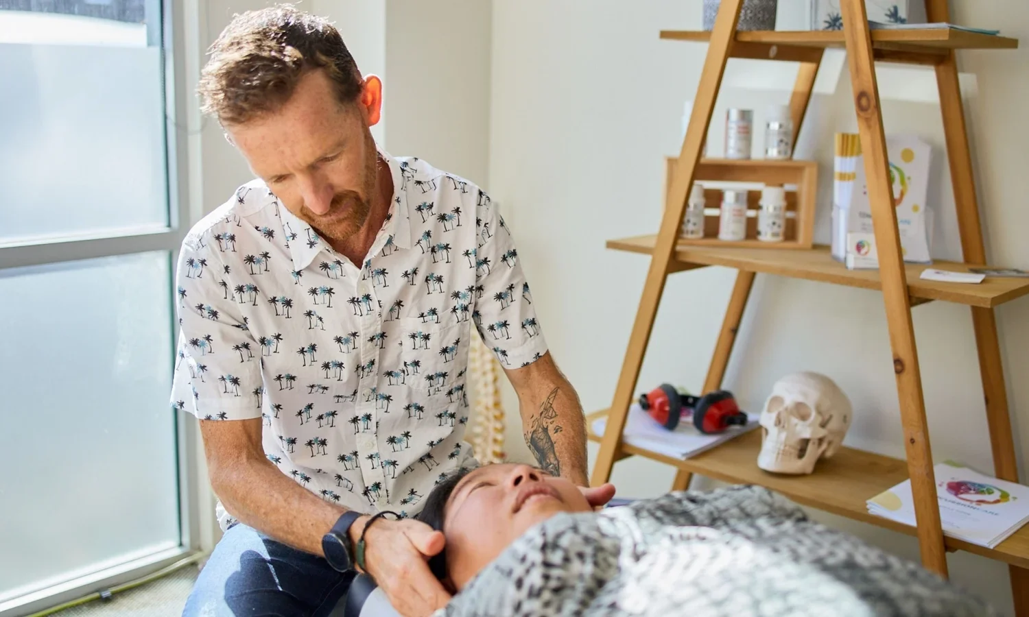 A man providing chiropractic care to a woman lying on a treatment table in a bright room with a wooden shelf holding bottles, papers, a skull model, and a drill. The man is wearing a tropical shirt and has a tattoo on his arm.