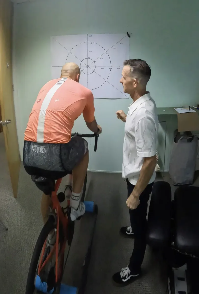 A man in a white shirt observing a person on a stationary bike, which is mounted on a blue platform, in an office with a dartboard on the wall.